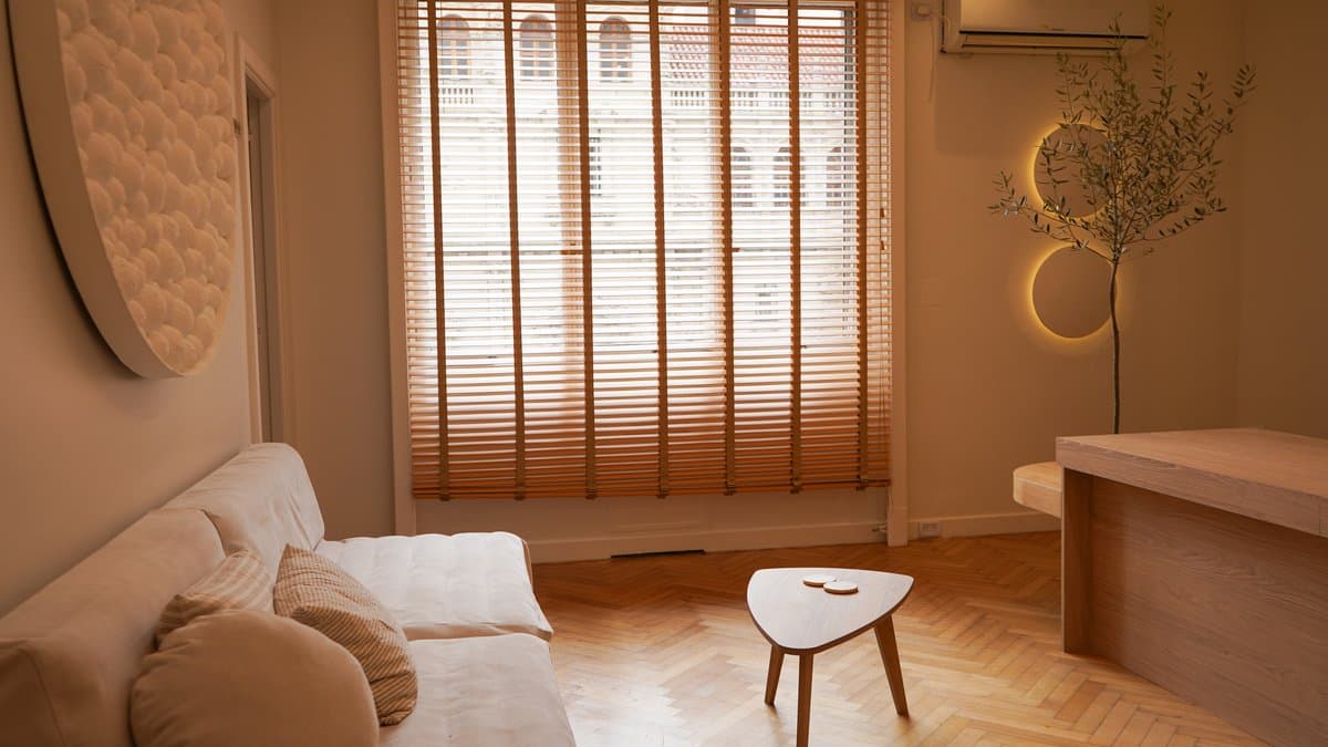 Sala de espera con sofá blanco, mesa ratona triangular y luz natural a través de persianas de madera